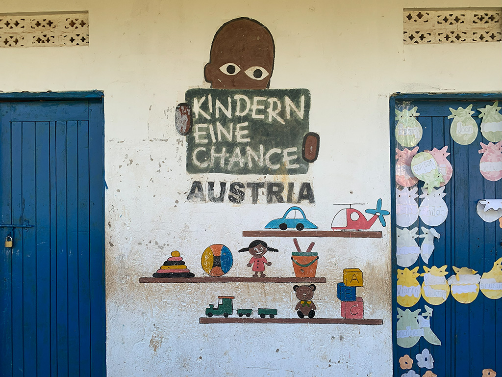 A mural on a wall shows a child holding a sign reading "Kindern eine Chance Austria," with painted shelves of toys and two blue doors on either side. Network Marketing Mastery