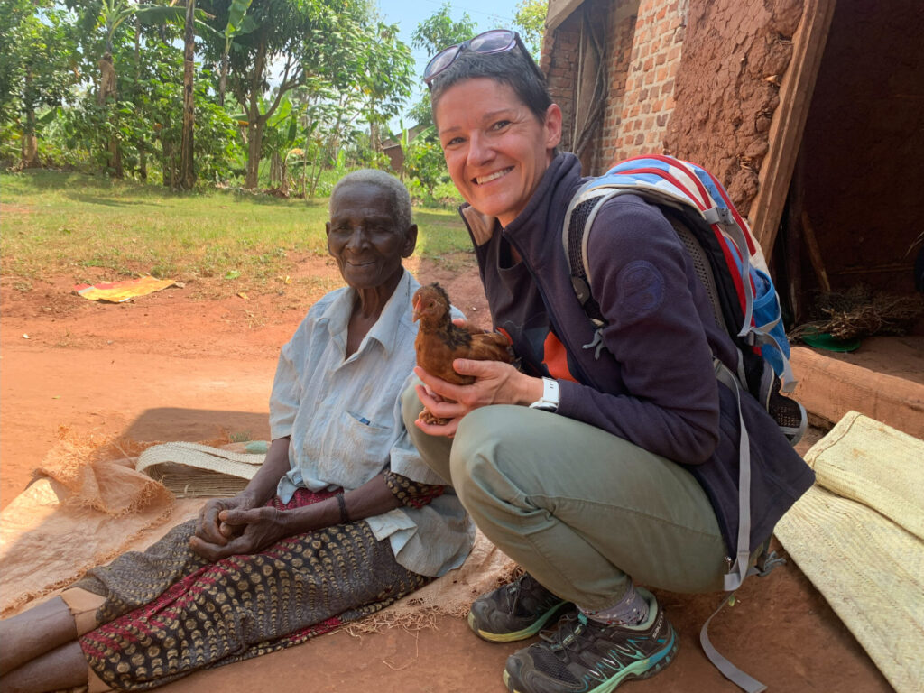 Two women sit outside a brick building; one holds a chicken while smiling at the camera. Lush greenery is visible in the background. Network Marketing Mastery