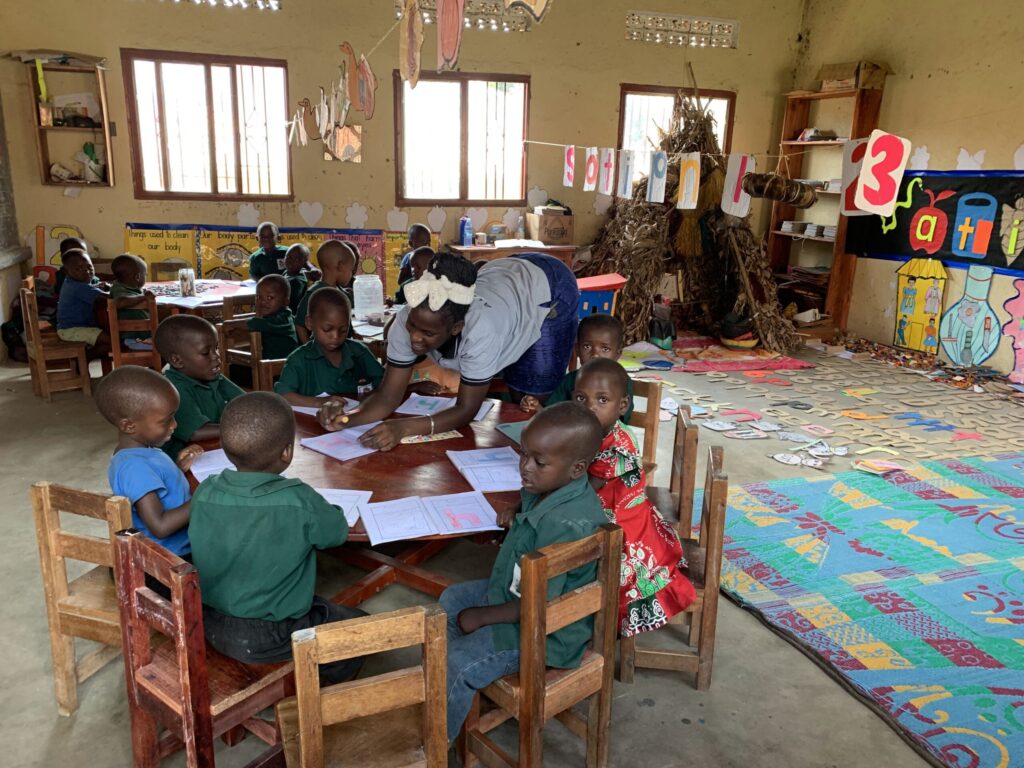 A teacher assists young children seated at round tables in a colorful classroom filled with learning materials and artwork. Network Marketing Mastery